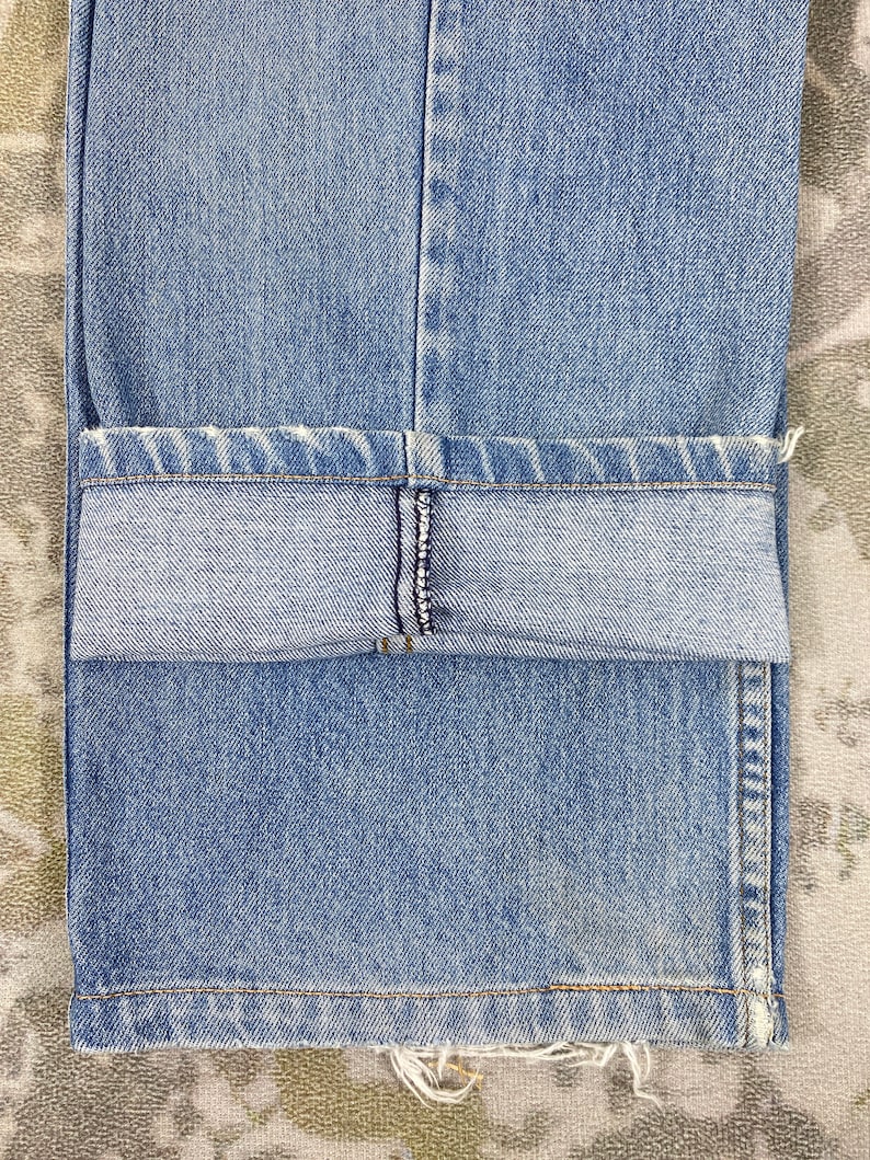 K&ouml;nnte beinhalten: Nahaufnahme einer hellblauen Jeans mit umgeschlagenem Saum. Die Jeans hat ein klassisches F&uuml;nf-Taschen-Design und einen leicht ausgefransten Saum. Der Stoff weist subtile Ausbleichungen und Abnutzungserscheinungen auf, was auf einen Vintage- oder beliebten Stil hindeutet.