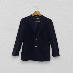 May include: Navy blue blazer with gold-colored buttons. The blazer has a classic two-button closure, notched lapels, and two front pockets. The garment is hanging on a wooden hanger against a white textured wall.