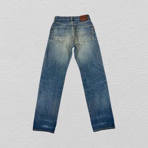 May include: Faded blue denim jeans with a classic five-pocket design. The jeans have a straight leg cut and a worn appearance, with a brown leather patch on the back waistband. The jeans are a size 32x32.