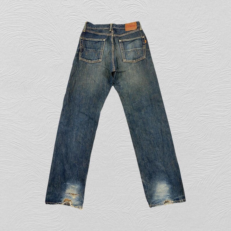 May include: Back view of blue denim jeans with a worn appearance. The jeans have a classic five-pocket design, with visible wear and tear at the bottom hems. A brown leather patch is on the back waistband.