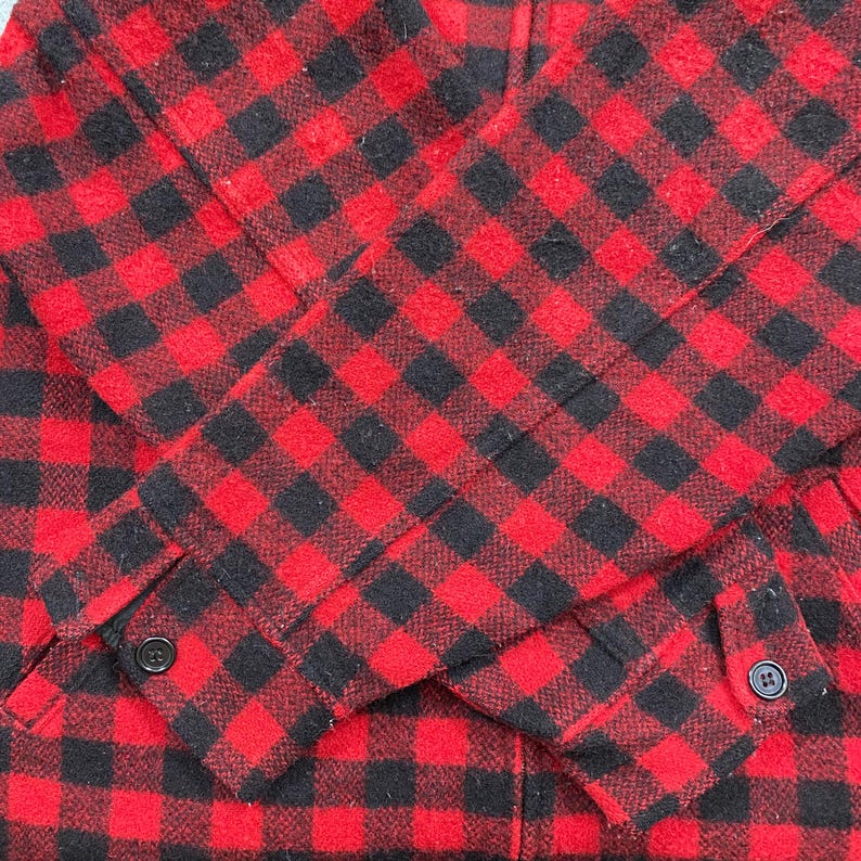 Op de afbeelding: Close-up van een rood-zwarte geruite jas. De jas heeft een klassiek ruitpatroon, zichtbaar op de mouwen en voorkant. De mouwen hebben manchetten met knopen. De stof lijkt een warm, geweven materiaal te zijn.