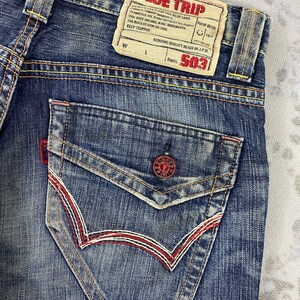 May include: Blue denim jeans featuring a back pocket with a red button closure and decorative red and white stitching. A beige leather-like patch at the waistband displays the text BLUE TRIP, RAW BLUE, and 503 with various printed details.
