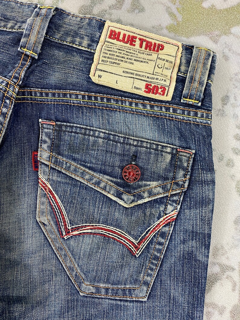May include: Blue denim jeans featuring a back pocket with a red button closure and decorative red and white stitching. A beige leather-like patch at the waistband displays the text BLUE TRIP, RAW BLUE, and 503 with various printed details.