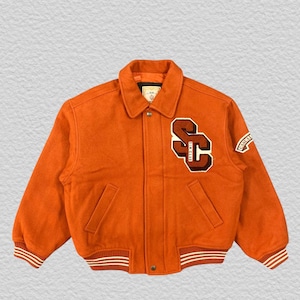 Puede incluir: Chaqueta universitaria naranja con cierre de botones, con un gran parche "SC" en marrón y blanco en el pecho. La chaqueta tiene cuello, dos bolsillos delanteros y puños y dobladillo a rayas. Un pequeño parche en la manga dice "SIM CLUB".