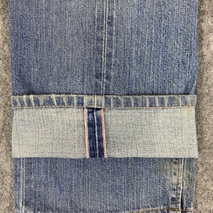May include: Close-up of blue denim jeans with a folded cuff. The jeans have a classic five-pocket design and a faded wash. The cuff reveals a red and white selvedge detail. The fabric is textured and shows signs of wear.