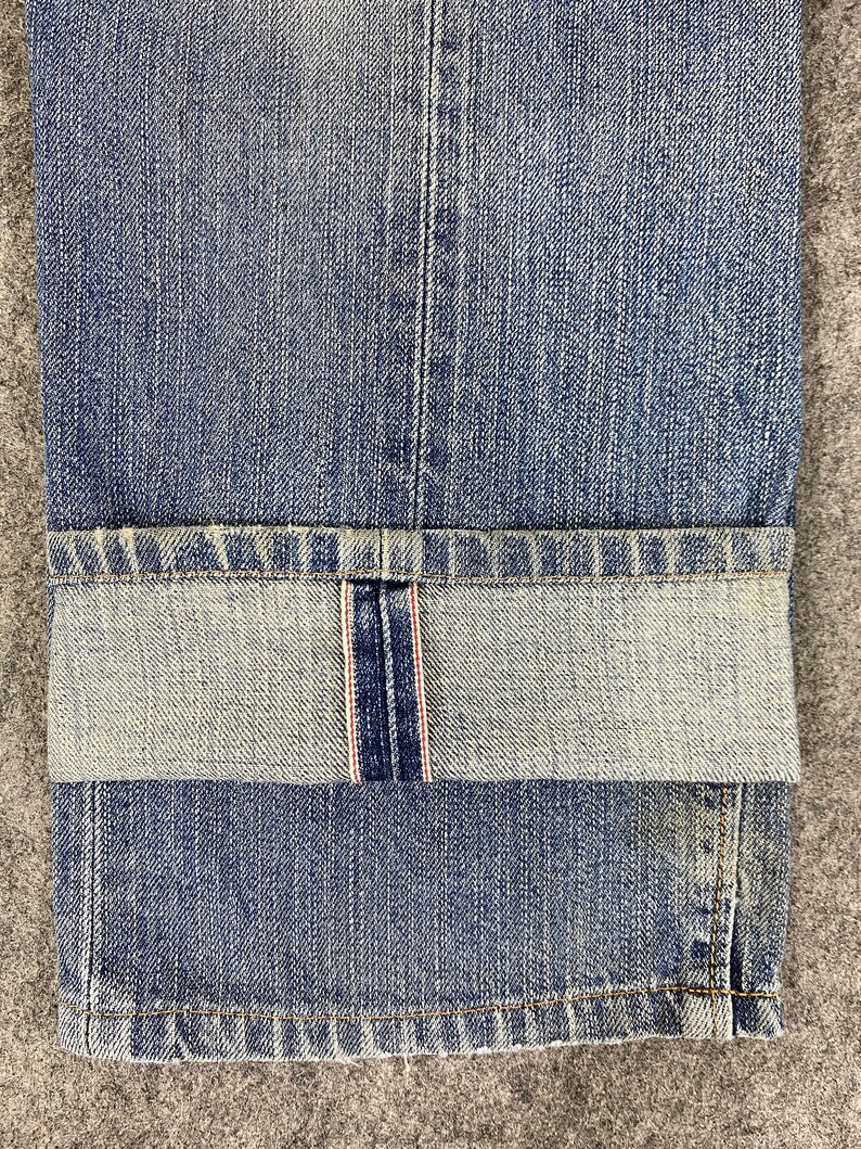 May include: Close-up of blue denim jeans with a folded cuff. The jeans have a classic five-pocket design and a faded wash. The cuff reveals a red and white selvedge detail. The fabric is textured and shows signs of wear.