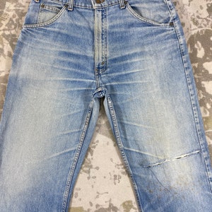 K&ouml;nnte beinhalten: Hellblaue Jeans aus Denim mit verwaschenem Look und abgenutztem Aussehen. Die Jeans haben ein klassisches F&uuml;nf-Taschen-Design, einen Knopf- und Rei&szlig;verschluss und einen abgenutzten Flicken am Knie. Die Jeans liegen flach auf einer gemusterten Oberfl&auml;che.