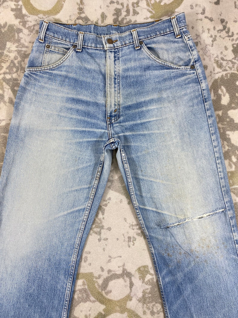 K&ouml;nnte beinhalten: Hellblaue Jeans aus Denim mit verwaschenem Look und abgenutztem Aussehen. Die Jeans haben ein klassisches F&uuml;nf-Taschen-Design, einen Knopf- und Rei&szlig;verschluss und einen abgenutzten Flicken am Knie. Die Jeans liegen flach auf einer gemusterten Oberfl&auml;che.