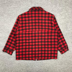 Op de afbeelding: Een rood-zwarte geruite jas met lange mouwen. De jas heeft een knoopsluiting aan de voorkant, een kraag en manchetten met knopen. De stof heeft een klassiek buffelruitpatroon. De jas is gemaakt van een dik, warm materiaal.