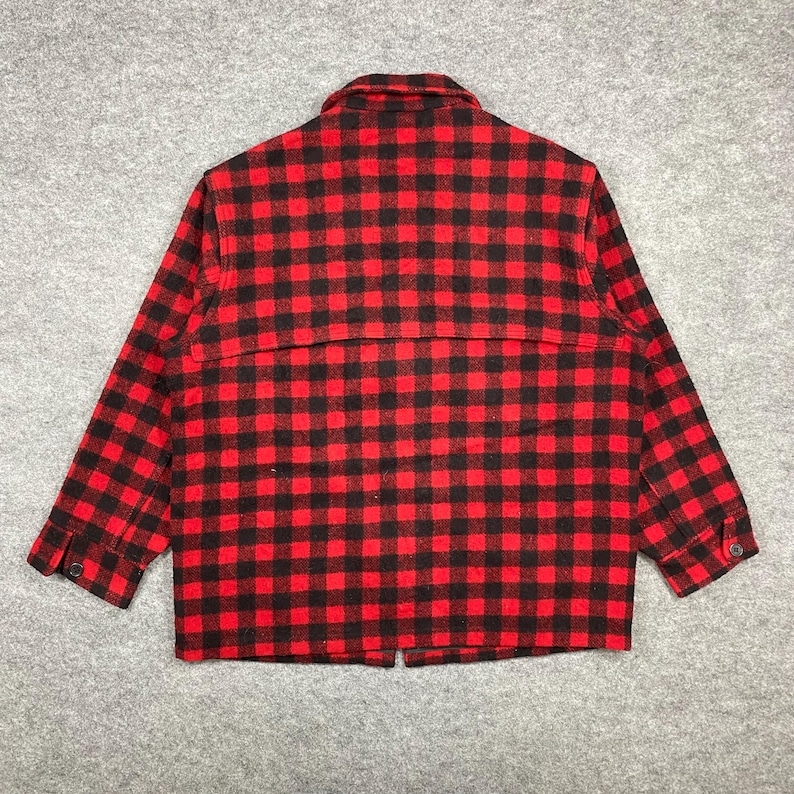 Op de afbeelding: Een rood-zwarte geruite jas met lange mouwen. De jas heeft een knoopsluiting aan de voorkant, een kraag en manchetten met knopen. De stof heeft een klassiek buffelruitpatroon. De jas is gemaakt van een dik, warm materiaal.