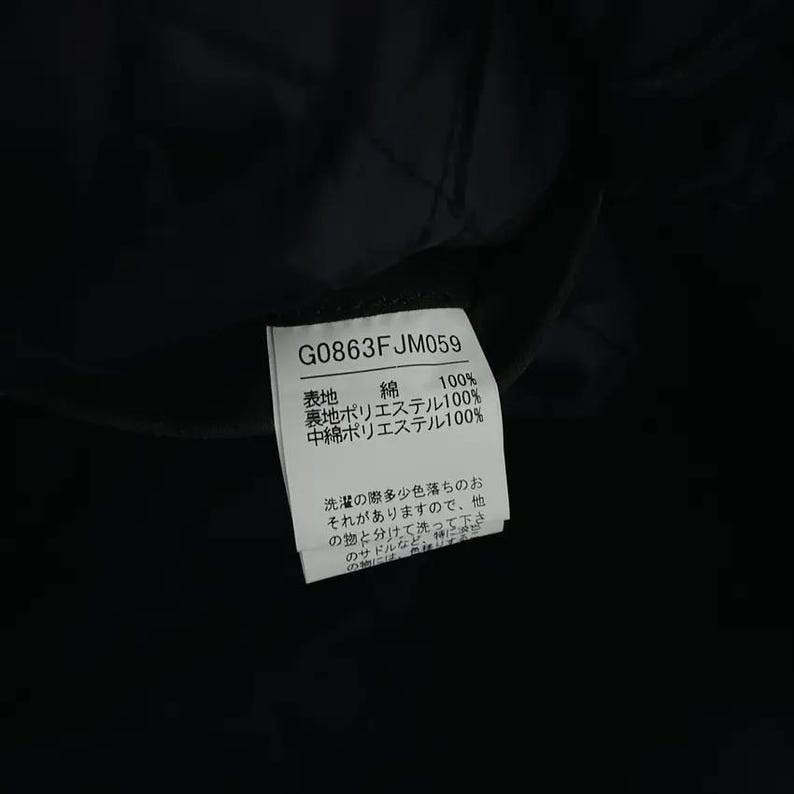 May include: A close-up of a black garment tag with the text "G0863FJM059". The tag lists fabric composition: 100% cotton, 100% polyester lining, and 100% polyester filling. Japanese text is also present.