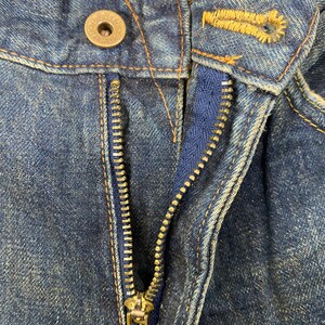 May include: Close-up of a pair of blue denim jeans, showing the button, buttonhole, and zipper. The zipper is open, revealing a blue fabric lining. The jeans have a worn, faded appearance, with visible stitching.