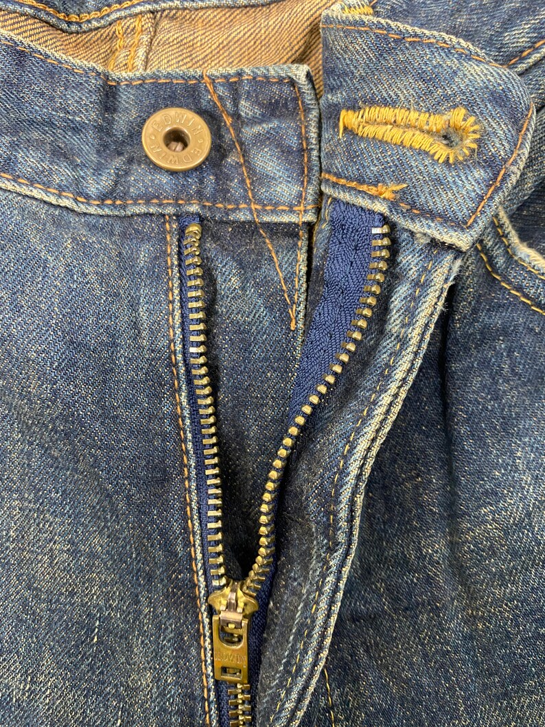 May include: Close-up of a pair of blue denim jeans, showing the button, buttonhole, and zipper. The zipper is open, revealing a blue fabric lining. The jeans have a worn, faded appearance, with visible stitching.