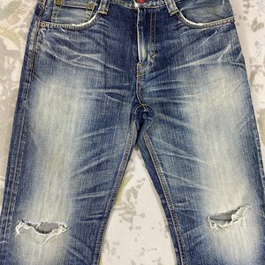 May include: Blue denim jeans featuring a distressed wash with faded whiskering patterns, a button fly, and two frayed holes at the knees. The interior waistband displays the number 503 in white text, and the garment includes copper-toned rivets.