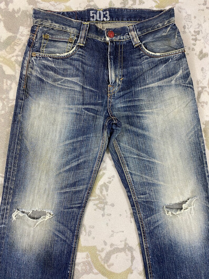 May include: Blue denim jeans featuring a distressed wash with faded whiskering patterns, a button fly, and two frayed holes at the knees. The interior waistband displays the number 503 in white text, and the garment includes copper-toned rivets.