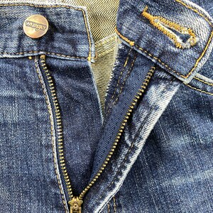 May include: Close-up of a pair of blue denim jeans with a brass zipper and a button with the word "Wrangler" on it.