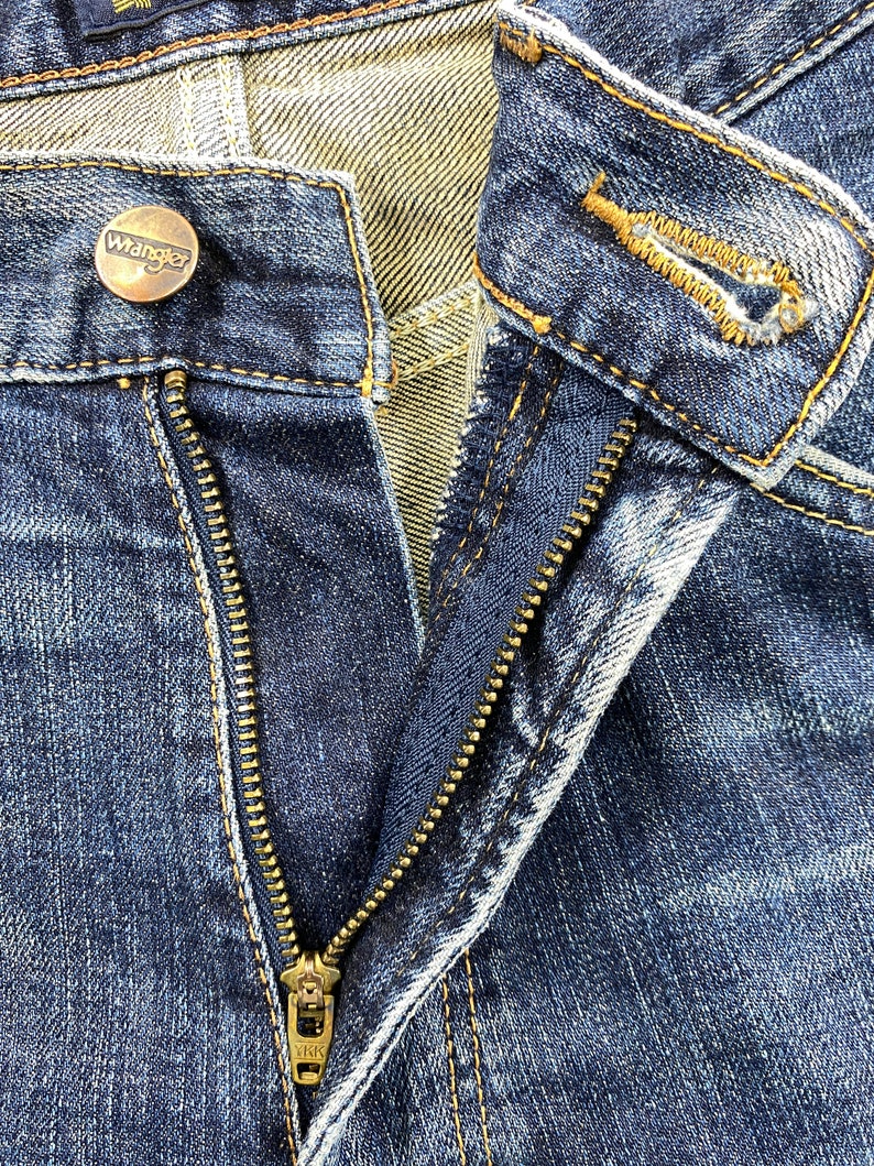 May include: Close-up of a pair of blue denim jeans with a brass zipper and a button with the word "Wrangler" on it.