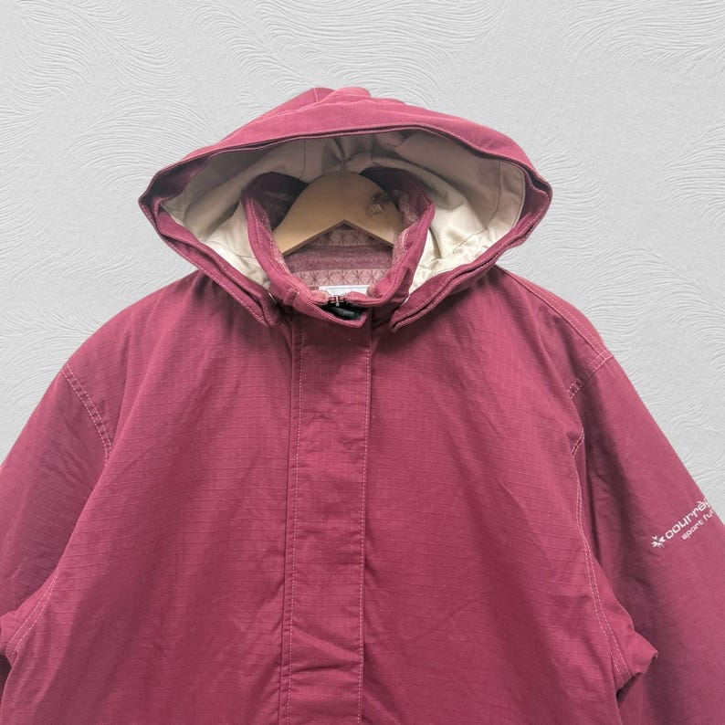 May include: A burgundy hooded jacket with a beige interior lining. The jacket has a full-length zipper and a high collar. The hood is adjustable and the jacket has a small logo on the sleeve. The jacket is made of a durable, weather-resistant material.