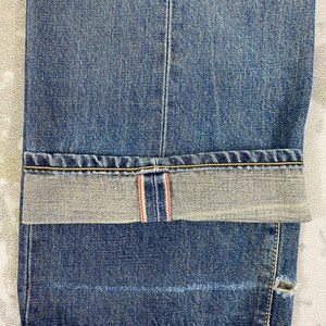 May include: Close-up of blue denim jeans with a folded cuff. The jeans have a classic five-pocket design, with visible stitching in yellow thread. The cuff reveals a red and white selvedge detail, and a small tear is visible near the hem.