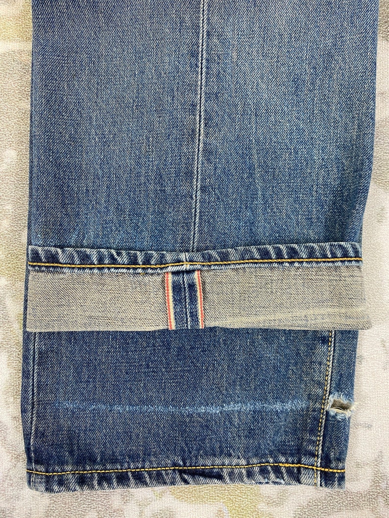 May include: Close-up of blue denim jeans with a folded cuff. The jeans have a classic five-pocket design, with visible stitching in yellow thread. The cuff reveals a red and white selvedge detail, and a small tear is visible near the hem.