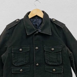 May include: A dark green military-style jacket with four front pockets and decorative buttons. The jacket has a collared neckline and shoulder epaulets. It is hanging on a wooden hanger.