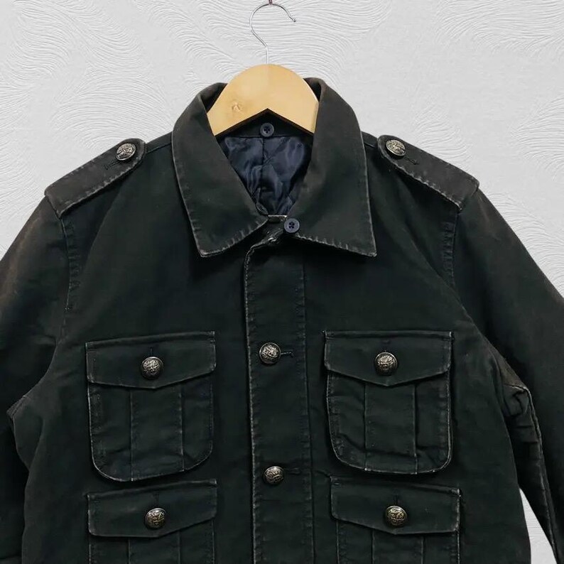 May include: A dark green military-style jacket with four front pockets and decorative buttons. The jacket has a collared neckline and shoulder epaulets. It is hanging on a wooden hanger.