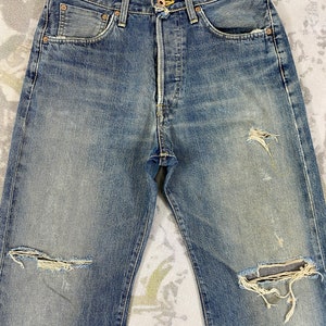 May include: Faded blue denim jeans with distressed details, including ripped knees and worn patches. The jeans feature classic five-pocket styling, a button fly, and copper rivets. The fabric shows signs of wear and fading, giving them a vintage look.