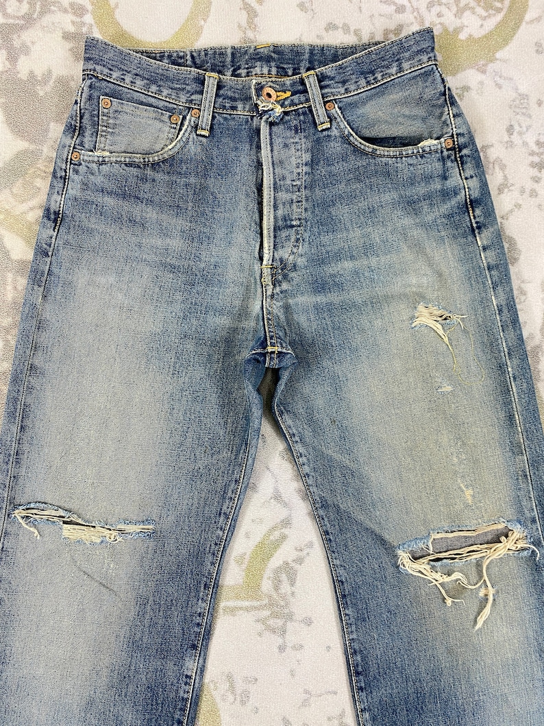 May include: Faded blue denim jeans with distressed details, including ripped knees and worn patches. The jeans feature classic five-pocket styling, a button fly, and copper rivets. The fabric shows signs of wear and fading, giving them a vintage look.