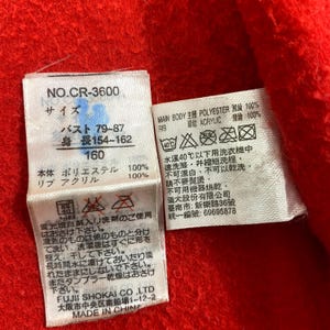K&ouml;nnte beinhalten: Nahaufnahme von wei&szlig;en Stoffetiketten mit japanischem Text und Pflegehinweisen, befestigt an einem roten Stoff. Die Etiketten enthalten Informationen zu Gr&ouml;&szlig;e, Material und Waschanleitung.
