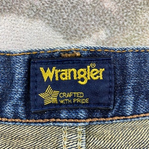 May include: Close-up of a blue denim jean waistband with a black Wrangler label. The label has a gold star with the words "Crafted with Pride" underneath.