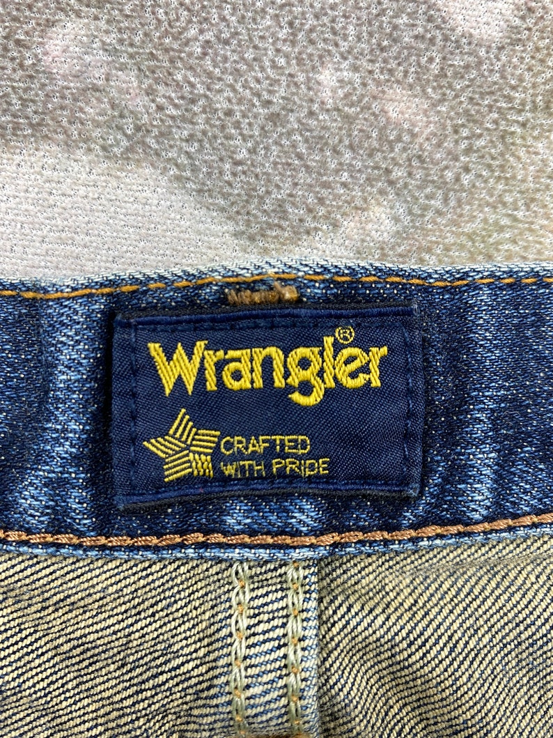 May include: Close-up of a blue denim jean waistband with a black Wrangler label. The label has a gold star with the words "Crafted with Pride" underneath.