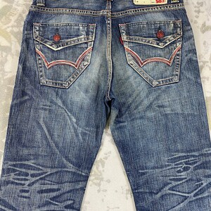 May include: Blue denim jeans featuring a faded wash with a subtle horizontal pattern. The back includes two flap pockets with red button closures and red stitching, along with a tan leather patch labeled BLUE TRIP 503 at the waistband.