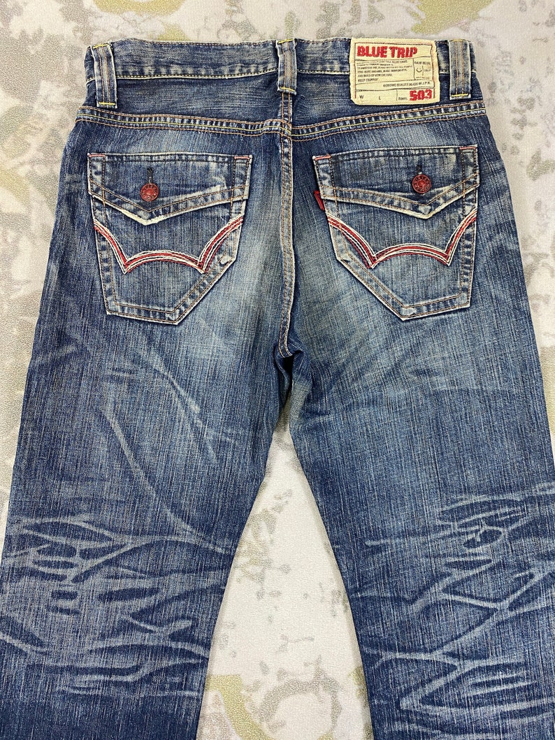 May include: Blue denim jeans featuring a faded wash with a subtle horizontal pattern. The back includes two flap pockets with red button closures and red stitching, along with a tan leather patch labeled BLUE TRIP 503 at the waistband.