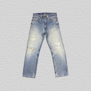 May include: Light blue denim jeans featuring a classic five-pocket design, a button fly closure, and a distressed finish with a noticeable tear on the left knee area. The fabric displays a faded wash pattern throughout the legs and hem.