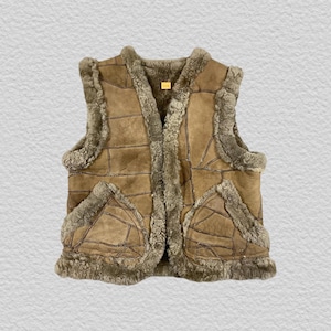 Op de afbeelding: Een beige suède vest met een patchworkontwerp en zichtbare donkerbruine stiksels. Het kledingstuk is afgewerkt met dikke, getextureerde grijsbruine schapenvacht langs de opening aan de voorzijde, de armsgaten en de zoom, inclusief twee zakken.