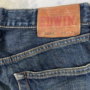 May include: Close-up of dark blue denim jeans with visible stitching and a brown leather patch. The patch features the brand name "EDWIN" and the text "505 X 30 34". The jeans are a classic style.