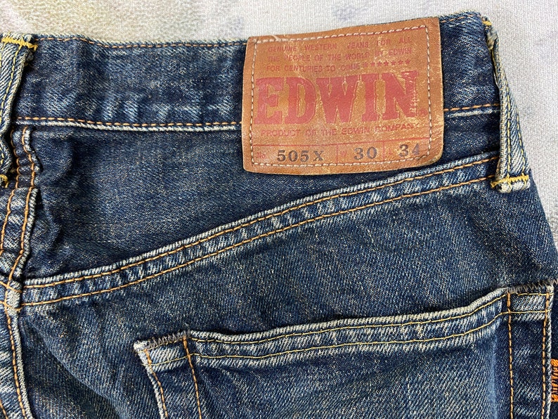 May include: Close-up of dark blue denim jeans with visible stitching and a brown leather patch. The patch features the brand name "EDWIN" and the text "505 X 30 34". The jeans are a classic style.