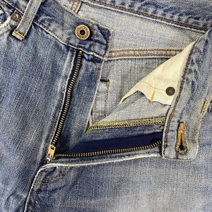 May include: Close-up of blue denim jeans with a partially unzipped zipper. The jeans show signs of wear and fading, with visible stitching and a button closure. The denim has a classic, worn look.