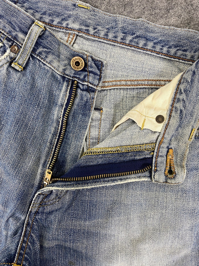 May include: Close-up of blue denim jeans with a partially unzipped zipper. The jeans show signs of wear and fading, with visible stitching and a button closure. The denim has a classic, worn look.