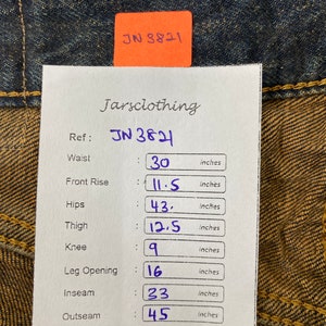May include: Close-up of blue denim jeans with a paper tag showing measurements. The tag reads "Jarsclothing" and "JN 3821". Waist: 30 inches, Front Rise: 11.5 inches, Hips: 43 inches, Thigh: 12.5 inches, Knee: 9 inches, Leg Opening: 16 inches, Inseam: 23 inches, Outseam: 45 inches.