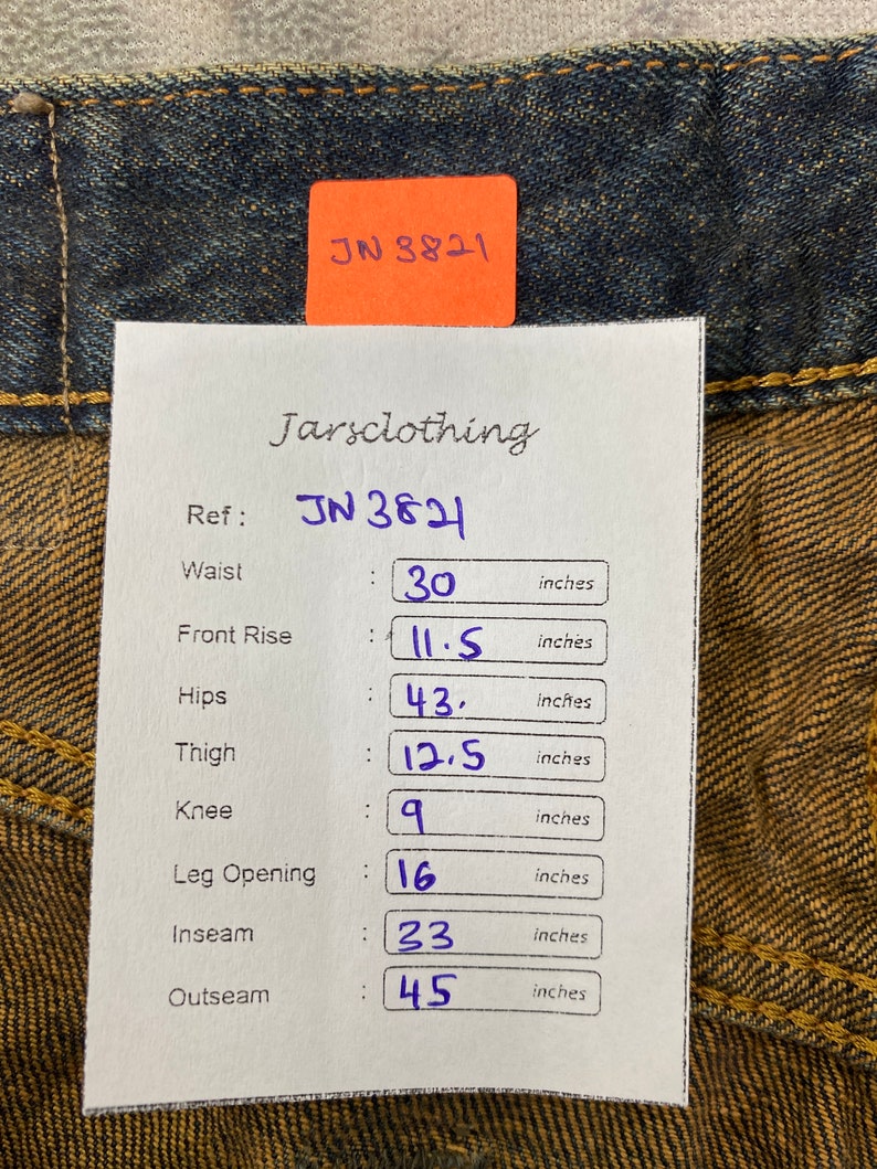 May include: Close-up of blue denim jeans with a paper tag showing measurements. The tag reads "Jarsclothing" and "JN 3821". Waist: 30 inches, Front Rise: 11.5 inches, Hips: 43 inches, Thigh: 12.5 inches, Knee: 9 inches, Leg Opening: 16 inches, Inseam: 23 inches, Outseam: 45 inches.
