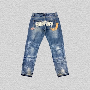 May include: Blue denim jeans featuring a faded wash, a back pocket with the text SURF UP! in white lettering, a small oval patch on the left side, and a yellow and blue plaid fabric accent on the right back pocket area with frayed hems.