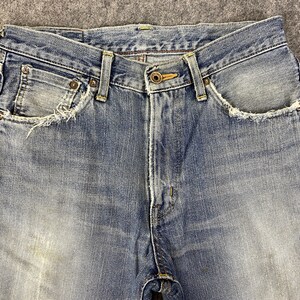 May include: Close-up of light blue denim jeans. The jeans have a button closure, belt loops, and front pockets with distressed edges. The denim has a faded appearance.