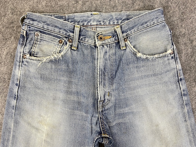 May include: Close-up of light blue denim jeans. The jeans have a button closure, belt loops, and front pockets with distressed edges. The denim has a faded appearance.
