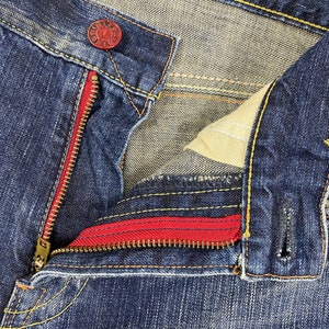 May include: Dark blue denim jeans featuring a red zipper tape, a brass-toned metal zipper, and a red metal button at the waist. The garment is constructed with contrasting yellow stitching along the seams and includes a beige interior pocket lining.