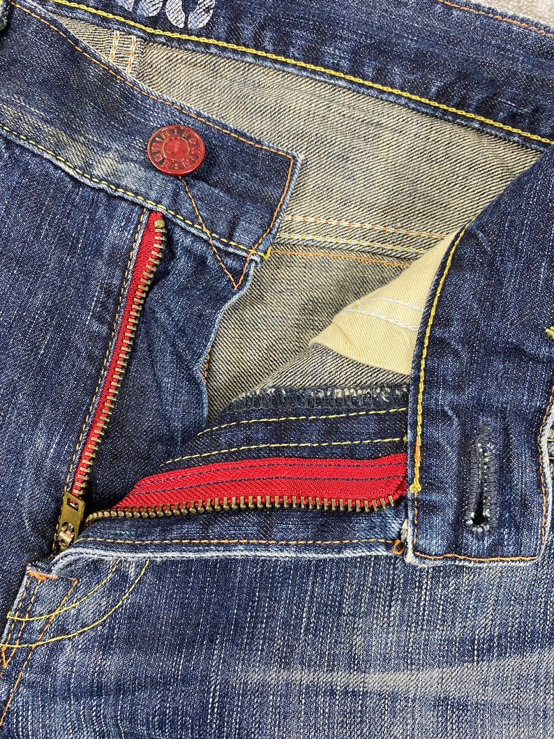 May include: Dark blue denim jeans featuring a red zipper tape, a brass-toned metal zipper, and a red metal button at the waist. The garment is constructed with contrasting yellow stitching along the seams and includes a beige interior pocket lining.