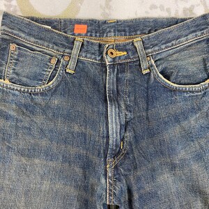 May include: Close-up of blue denim jeans. The jeans have a button and zipper closure, with rivets and pockets. The denim has a faded, worn look. The tag reads "JM 5543".