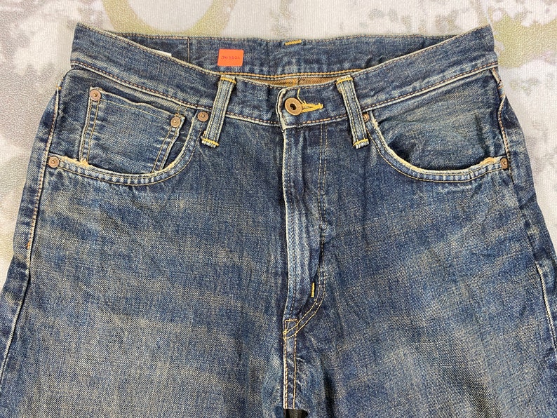 May include: Close-up of blue denim jeans. The jeans have a button and zipper closure, with rivets and pockets. The denim has a faded, worn look. The tag reads "JM 5543".