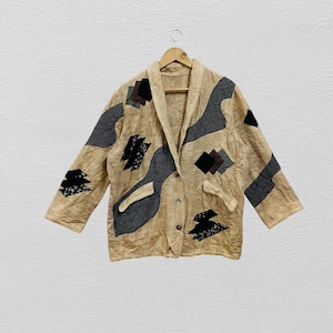 May include: A tan jacket with grey and black patchwork details. The jacket features a collared neckline, long sleeves, and a button-down front. The design incorporates geometric shapes and abstract patterns, creating a unique, artistic look. It is hanging on a wooden hanger.