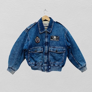 May include: A blue denim bomber jacket featuring a front zipper closure, two large flap pockets, and ribbed cuffs. The jacket displays a circular crest patch on the right chest and a rectangular patch on the left chest with the text MT-55 LOGX WORKOUT.
